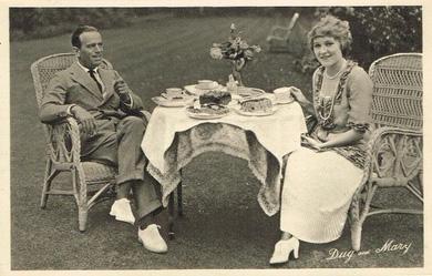 Douglas Fairbanks and Mary Pickford sit outside on wicker chairs either side of a small round table.